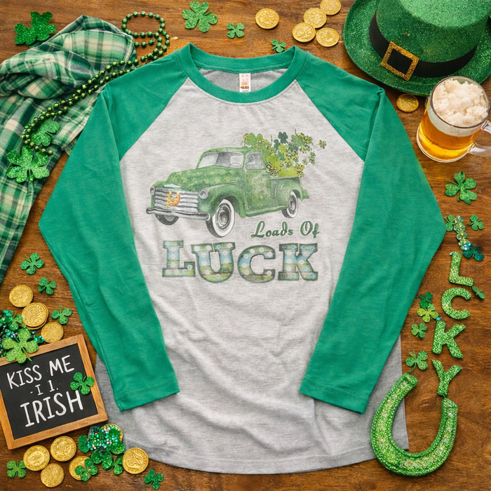 Green and gray baseball shirt with vintage truck design and 'Loads of Luck' text, surrounded by St. Patrick's Day decorations on a wooden surface.